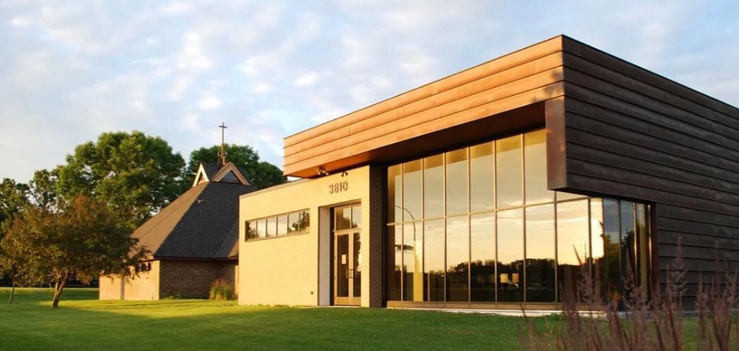 A historical adobe-style church with a cross, wooden accents, and bell. Surrounded by trees under a clear blue sky.