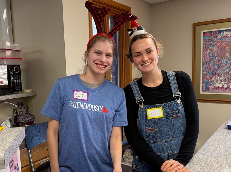 Two people wearing festive headbands smiling in a kitchen setting with name tags. Artwork hangs on the wall in the background.