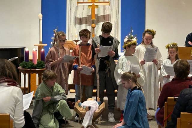 Children in costume perform a nativity scene inside a church, with lit candles and a cross in the background. People watch attentively.
