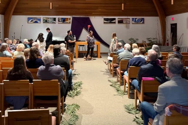A small gathering in a wooden hall. Several people stand while others sit on wooden chairs, surrounded by palm fronds on the floor.
