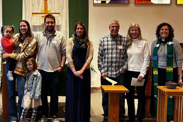 A group of people, including a child, stand indoors in front of a cross, likely in a church setting.