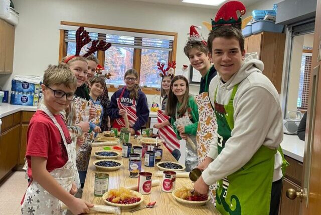 People and children, wearing festive headbands, prepare pies together around a table in a bright, cozy room with pantry items.