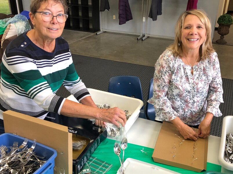 Two people organize cutlery at a table inside a room, with bins and boxes around. The setting appears casual and collaborative.