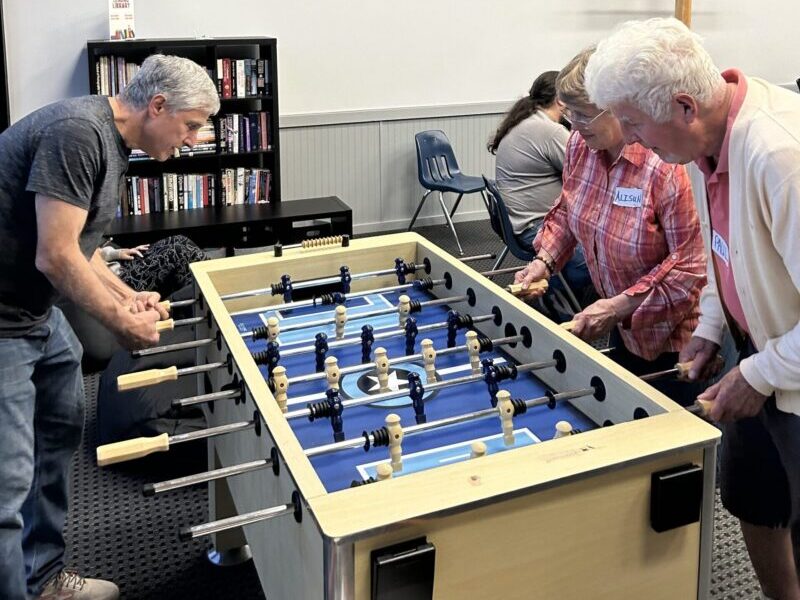 Three people play foosball in a room with origami birds on the wall, a bookshelf, and a wooden cross in view.