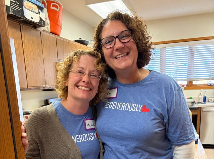 Two smiling people in a kitchen wearing matching blue shirts with a heart design. They have name tags and appear friendly and joyful.