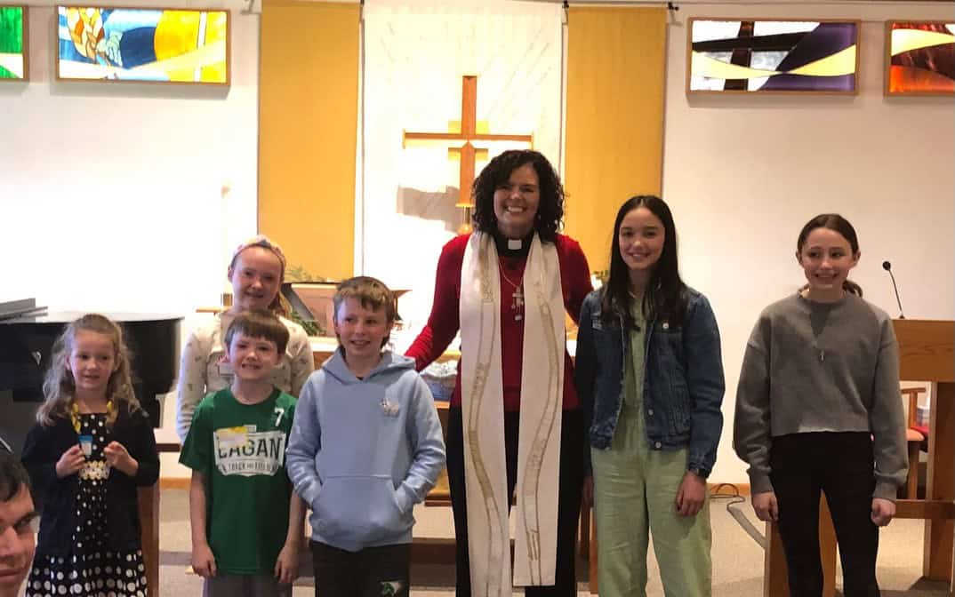 A person and six children stand together in a church, with colorful stained glass and a cross in the background.