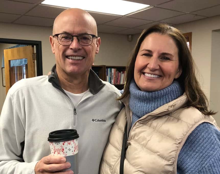 Two smiling people inside a room with bookshelves, one holding a coffee cup. Both are dressed in cozy sweaters and vests.