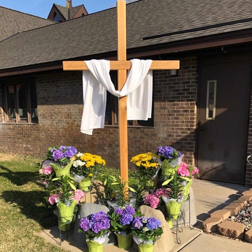 A wooden cross with a white cloth is surrounded by colorful flowers outside a brick building, capturing a serene spring scene.