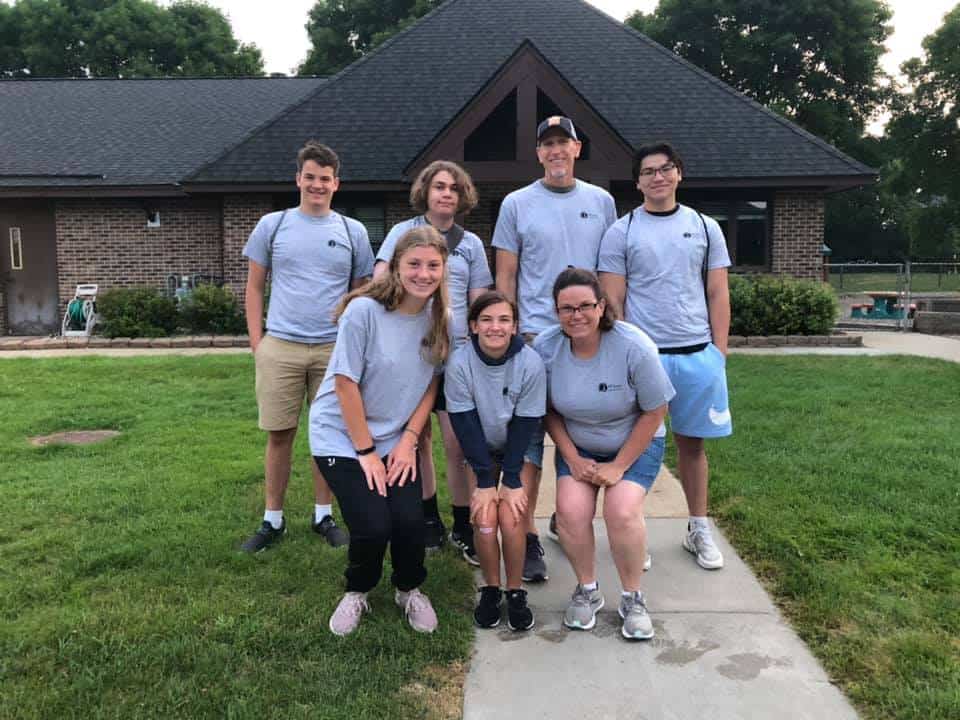 Seven people in matching gray shirts gather outdoors, smiling in front of a brick building with a triangular roof. Grassy area surrounds them.