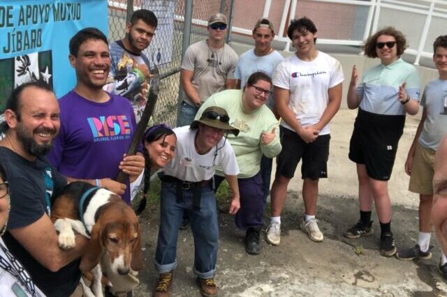 A group of people gathered outdoors, smiling and holding a dog, with a sign in the background. There are no recognizable landmarks.