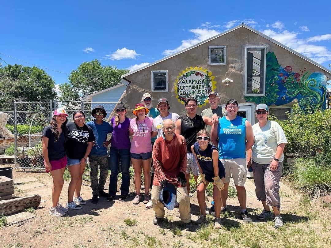 A group of people poses cheerfully outside the Alamosa Community Greenhouse, with vibrant murals and a clear blue sky above.