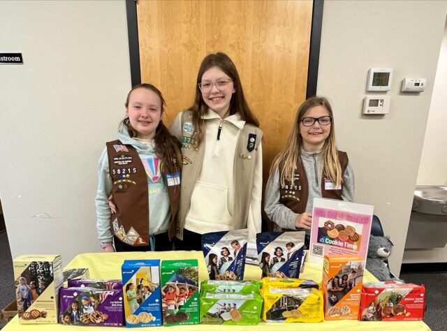 Three children in uniforms at a cookie sale table with various cookie boxes and a promotional sign for "Troop 57704."