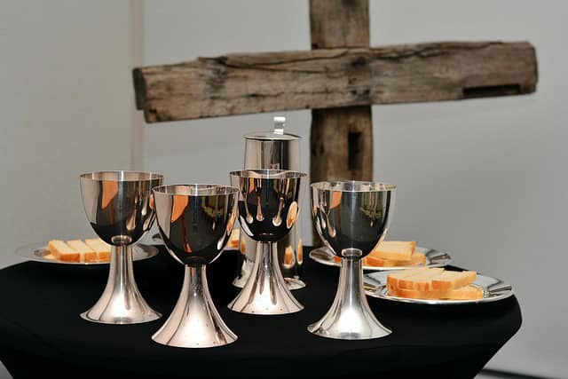 Communion setup with silver chalices and bread on a table. A wooden cross is prominently displayed in the background, suggesting a religious setting.