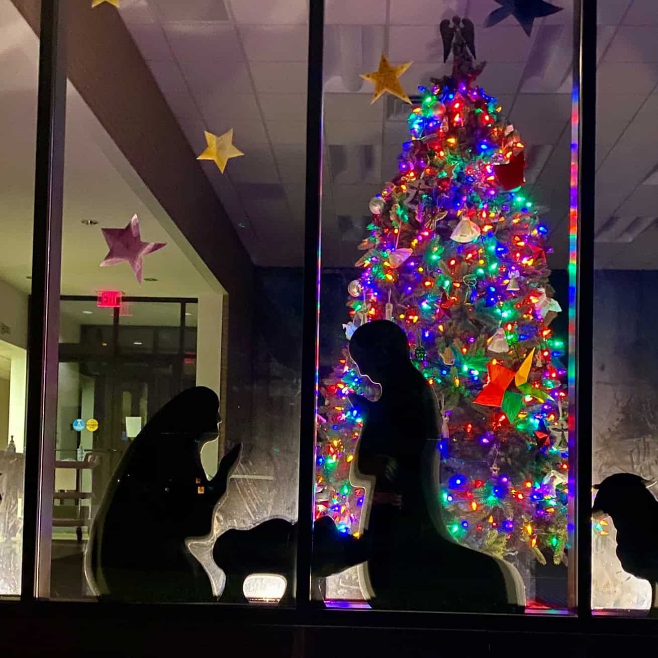 Silhouette figures next to a colorful, lit Christmas tree inside a building. Stars hang nearby, creating a festive holiday atmosphere.
