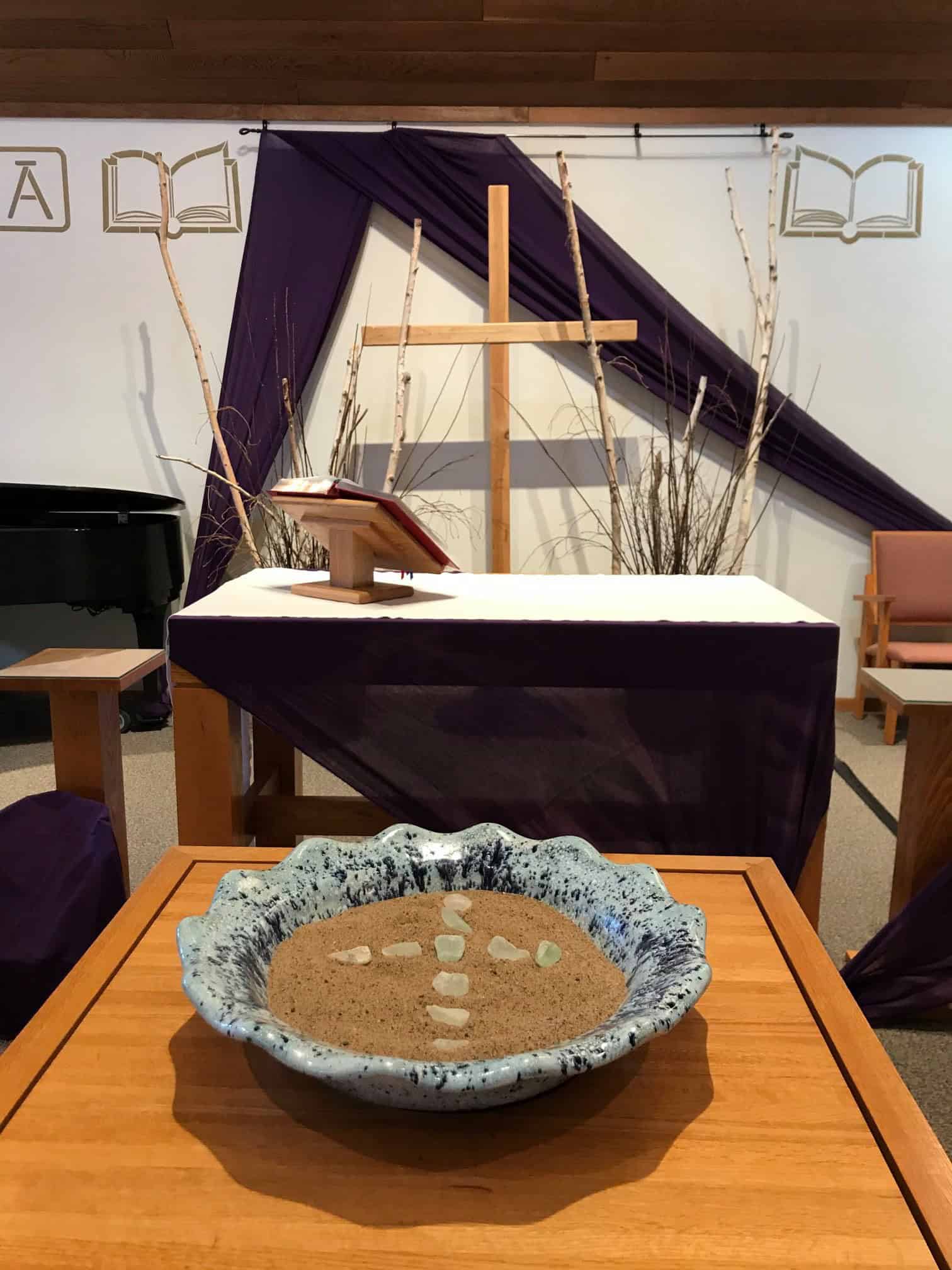 A church altar with a wooden cross, purple drapery, scriptures, sand bowl, and decorative sticks. Peaceful, spiritual setting with subtle decor.
