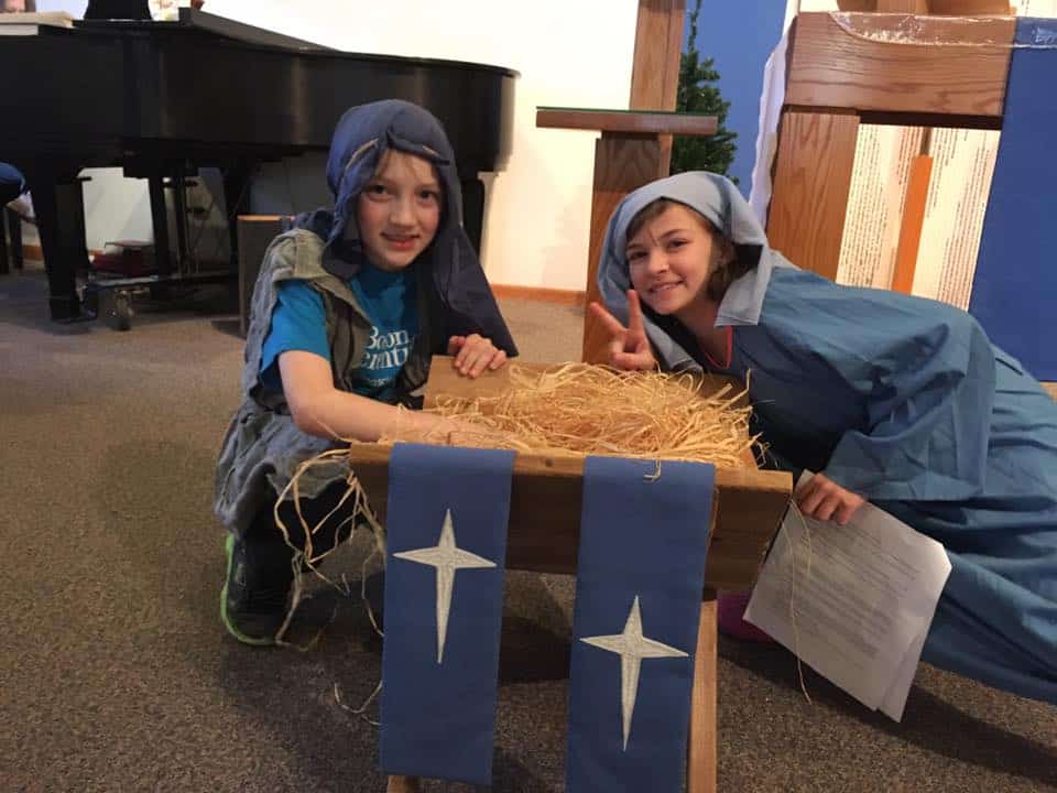Two children in nativity outfits pose by a manger with straw and blue cloth, inside a room with a piano.
