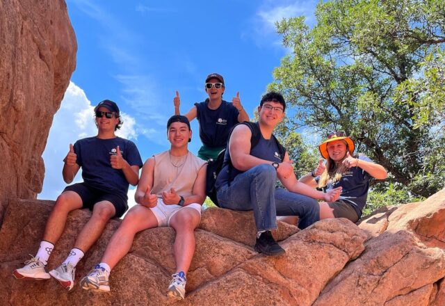 Five people sit on a large rock formation, enjoying a sunny day. They smile and give thumbs up, surrounded by trees and a clear sky.