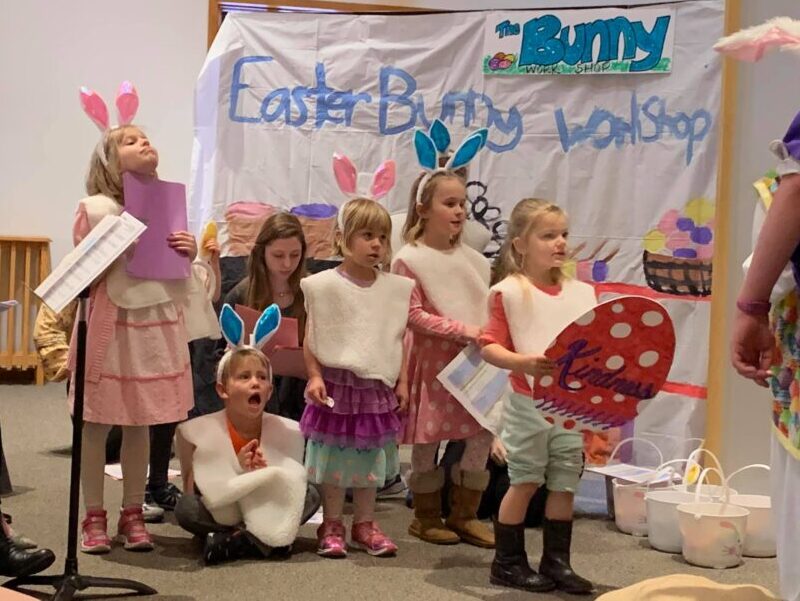 Children wearing bunny ears perform at "Easter Bunny Workshop" event. They hold signs and papers; adults assist in background.