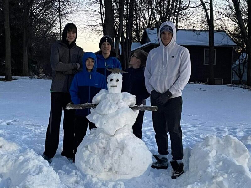 Five people standing around a snowman in a snowy landscape with trees and a house in the background.