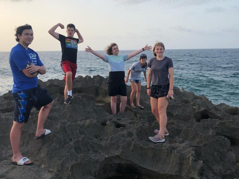 Five people stand on jagged rocks by the ocean, posing energetically. The sky is partly cloudy, and the sea appears calm.