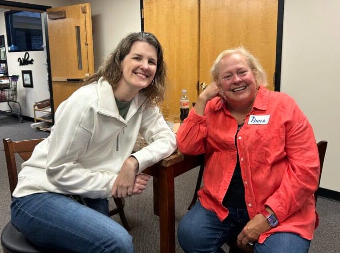 Two people seated and smiling indoors, with open doors in the background. A name tag is visible on one person. Office environment.
